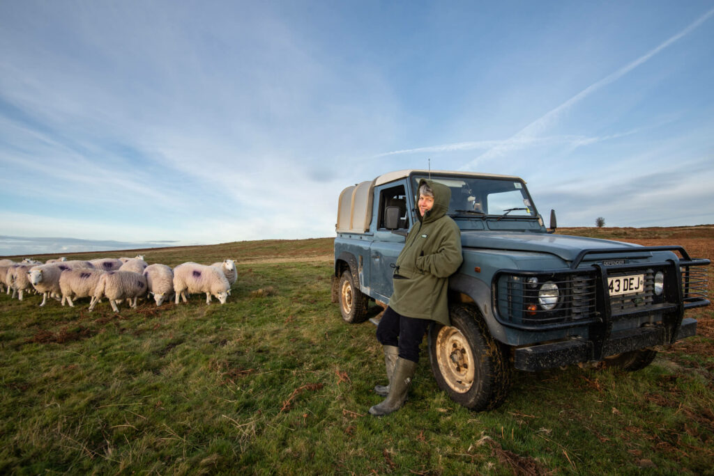 A female farmer leans against her Land Rover in the foreground with sheep in the background.